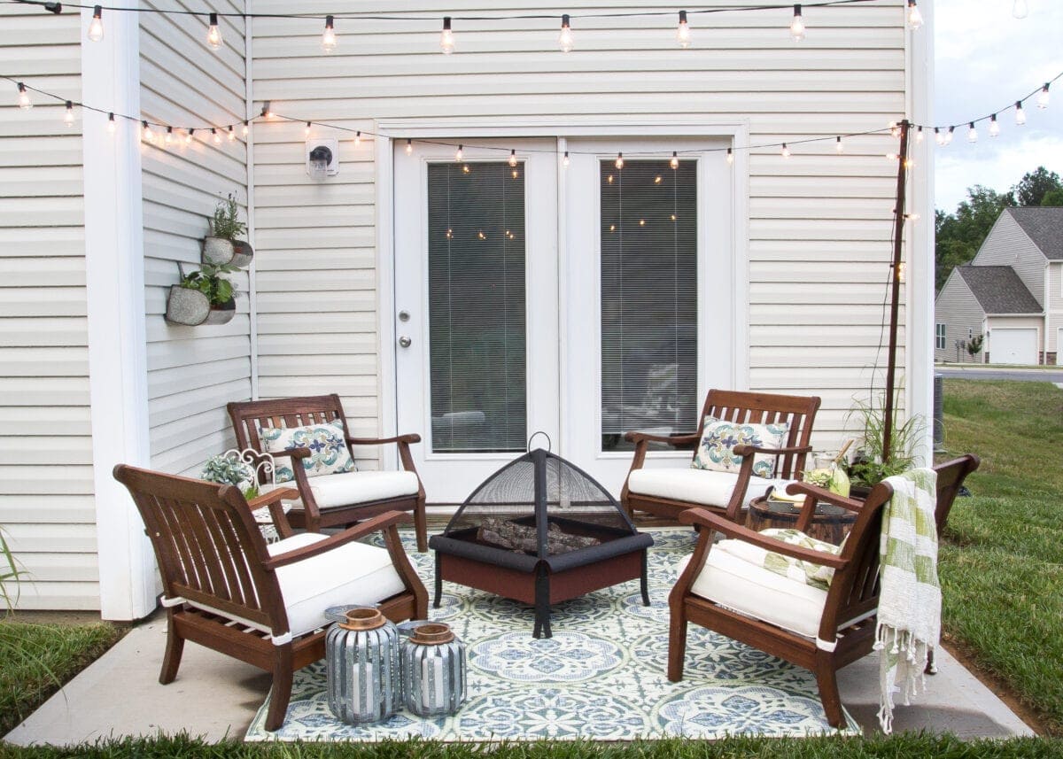 string lights hanging on small back patio lit up during day time wood chairs with white cushions around a fire pit