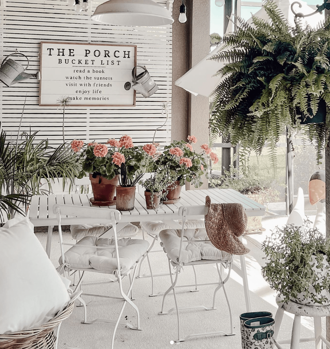 white patio furniture on porch with farmhouse style sign on wood slat wall fern potted plants on table pillows