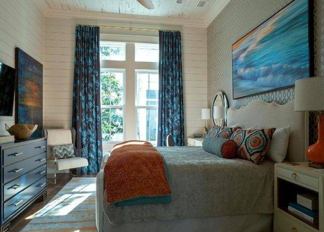 large art piece hangs from a wall clad in gold and blue trellis wallpaper over a tan wood and fabric bed complemented with orange and blue pillows complementing an orange and blue blanket. The bed is flanked by orange gourd lamps placed on grasscloth nightstands located under round mirrors.The bed sits on an orange and blue rug under a pecky cypress ceiling. White shiplap frames windows coved in long blue curtains.