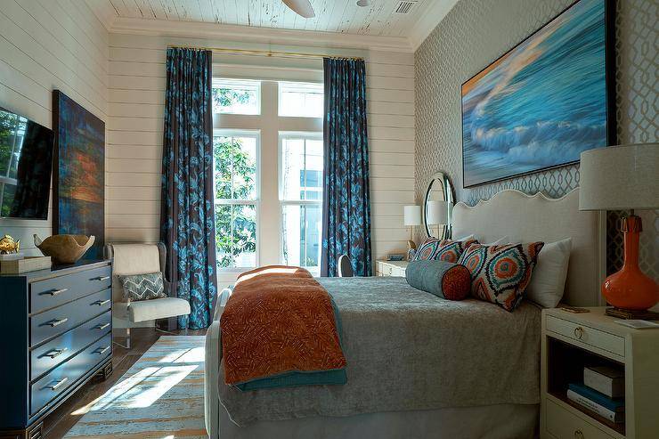 large art piece hangs from a wall clad in gold and blue trellis wallpaper over a tan wood and fabric bed complemented with orange and blue pillows complementing an orange and blue blanket. The bed is flanked by orange gourd lamps placed on grasscloth nightstands located under round mirrors.The bed sits on an orange and blue rug under a pecky cypress ceiling. White shiplap frames windows coved in long blue curtains.