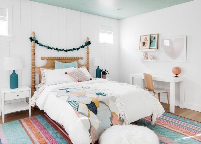 White nightstands accented with peacock blue bottle lamps are placed in front of a vertical shiplap wall on either side of a vintage spindle poster bed accented with a blue pom pom garland. The bed sits on a colorful rug beneath a blue painted ceiling. A white desk is located beneath a heart art piece and stacked white floating shelves.