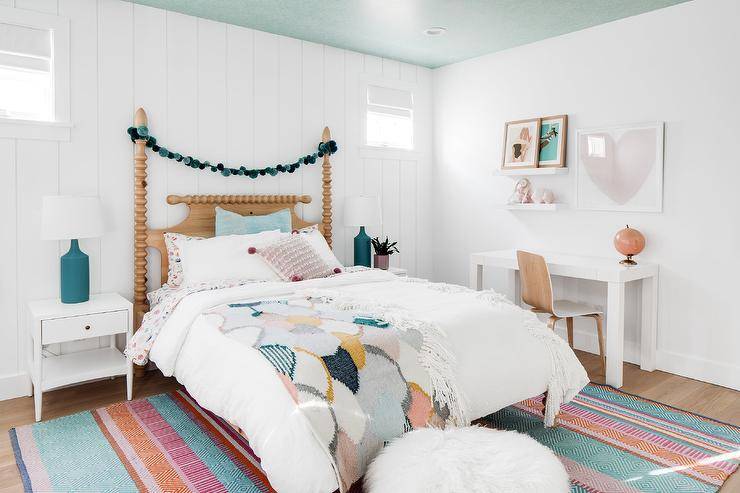 White nightstands accented with peacock blue bottle lamps are placed in front of a vertical shiplap wall on either side of a vintage spindle poster bed accented with a blue pom pom garland. The bed sits on a colorful rug beneath a blue painted ceiling. A white desk is located beneath a heart art piece and stacked white floating shelves.
