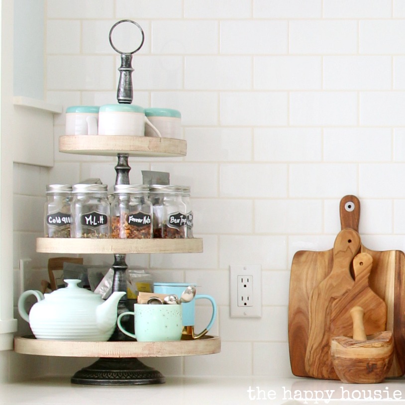 three tiered tray with spice jars on them mugs and teapot white subway tile background next to wood handle cutting boards