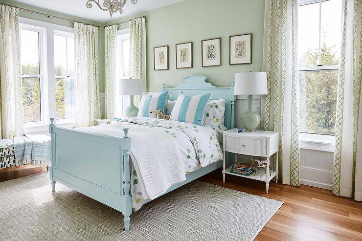 Blue and green farmhouse bedroom is furnished with a blue French wood bed dressed in white and green bedding complemented with blue striped pillows. The bed sits on a gray rug against a shiplap trim and beneath four botanical art pieces hung side by side from a green painted wall. White curved nightstand lit by a celadon green lamps flank the bed, as windows are covered with white and green lattice curtains.