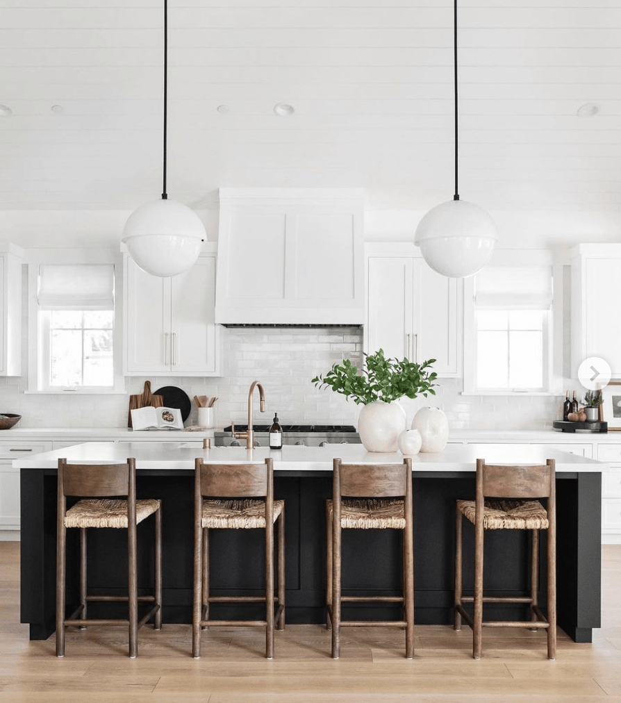 black island in white kitchen with hanging pendants cabinetry stools wicker and wood greenery on countertop