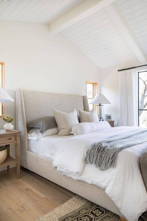 Bedroom features a plank vaulted ceiling over an oatmeal linen wingback bed with gray bedding, flanked by brown farmhouse nightstands, atop layered gray and tan rugs.