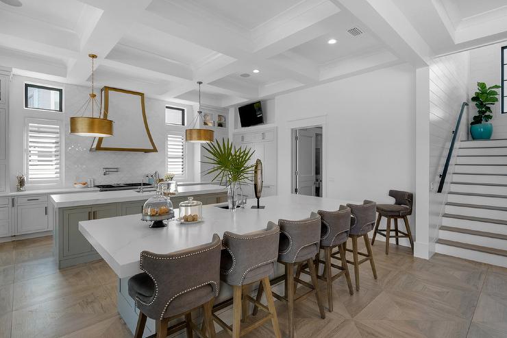 Brown stools sit on a parquet wood floor at a gray kitchen island finished with a prep sink and are positioned facing a matching gray island lit by gold drum pendants hung from a coffered ceiling.