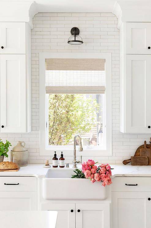 white wood shade window behind white farmhouse apron sink white shaker cabinets black wall sconce flowers in sink