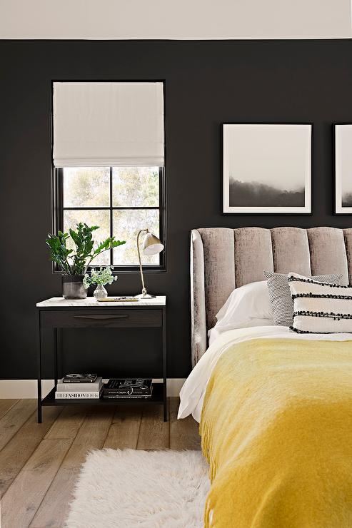Bedroom features a gray channel tufted wingback bed with a yellow throw blanket, a black and white marble top nightstand lit by a brass lamp and black walls.