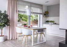 Gray roman shades and a pink curtain on big, glass windows in a modern kitchen and dining room interior with a wooden table and white chairs