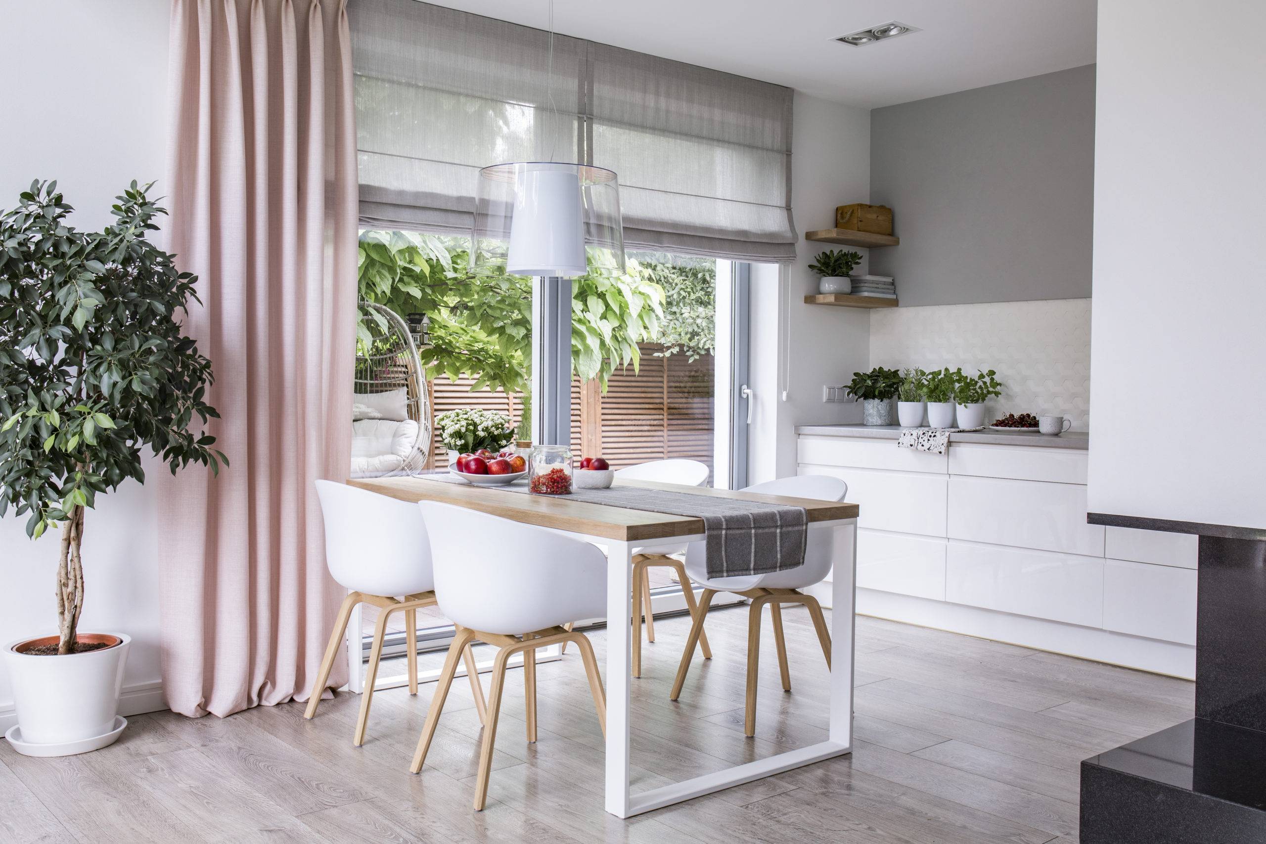 Gray roman shades and a pink curtain on big, glass windows in a modern kitchen and dining room interior with a wooden table and white chairs