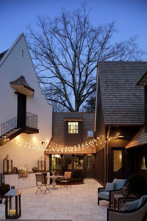 String lights and large outdoor candle lit lanterns illuminate two wood and iron cafe chairs sitting in front of a round marble cafe table sat atop tan slate floors and in front of an outdoor kitchen fitted with white flat faced cabinets and a stainless steel grill. In the corner two swoop arm patio chair separated by small square side table.