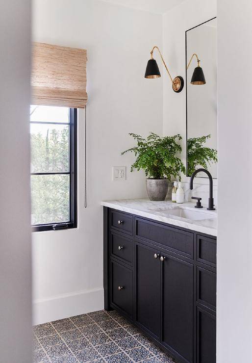 A black washstand sits on black and gray quatrefoil floor tiles and is topped with a white marble countertop finished with an oil rubbed bronze gooseneck faucet. A black metal framed vanity mirror is flanked by black and gold sconces.