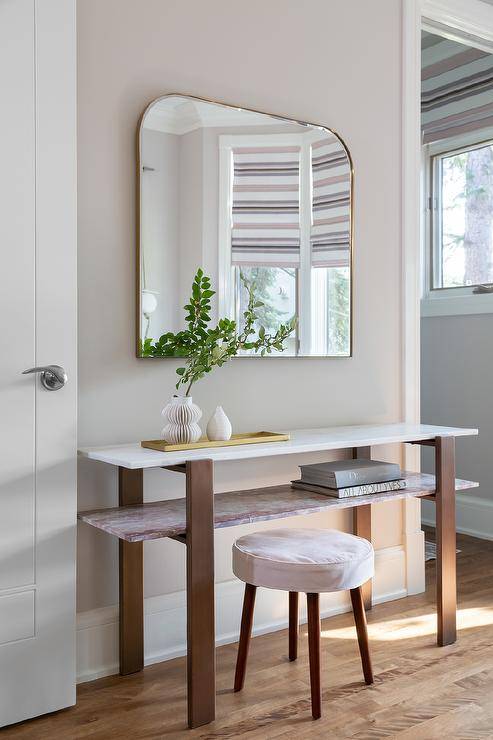 Entryway features a pink marble shelf console table under a mounted brass arch mirror with a white and brown wooden stool.