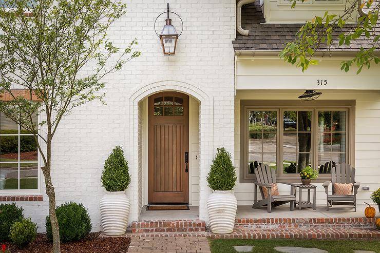 Cottage home with a white brick exterior, an arched entryway and a wood door accented by white rippled planters atop red brick floors. A carriage lantern lights the entryway over the arched entryway onto a porch that includes gray Adirondack chairs, a gray slatted outdoor table, and orange accent pillows.
