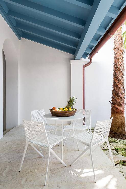 Covered patio with a blue plank ceiling furnished with a white outdoor table and modern white chairs surrounded by lush greenery and palm trees.