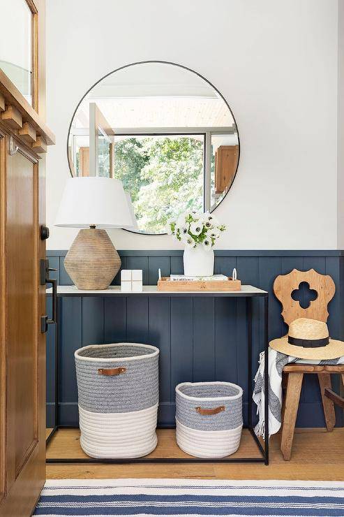 Cottage style foyer features a round black mirror on a white wall over blue vertical shiplap trim and a vintage reeded lamp atop an iron and marble console table with white and gray woven bins.