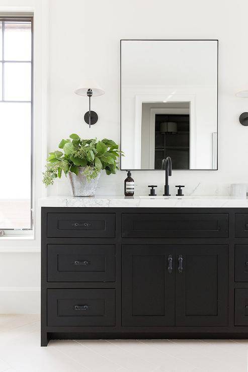 Black pulls on a black washstand vanity balanced with stunning white marble countertop and a matte black gooseneck faucet. A black metal framed mirror keeps the black and white theme with a sleek thin frame flanked by contemporary black and white sconces.