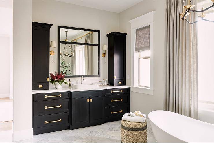 Brass knobs complement a black shaker washstand contrasted with a white quartz countertop completed with a vessel sink. The washstand is finished with black linen cabinets and is positioned beneath a black framed vanity mirror