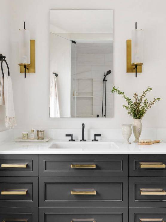 Brass and glass sconces flank a frameless vanity mirror mounted above a black washstand donning brass pulls and white marble countertop finished with a matte black gooseneck faucet.