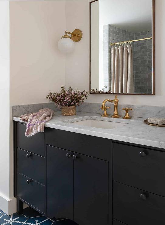 A brass and glass globe sconce lights a vanity mirror hung over a black sink vanity adorned with black hardware and a gray marble countertop finished with a brass hook and spout faucet. The washstand sits on blue hexagon floor tiles.