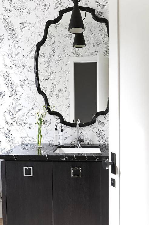 Chic black and white powder room features a glossy black mirror mounted on black and white wallpaper over a black wooden washstand topped with polished black marble and a sink with a polished nickel faucet.