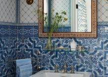 Mediterranean style bathroom features a gold Greek key mirror hung from a wall clad in mixed white and blue mosaic tiles between brass sconces and over a glass shelf. The shelf is fixed above a white porcelain pedestal sink vanity finished with a polished nickel faucet kit.