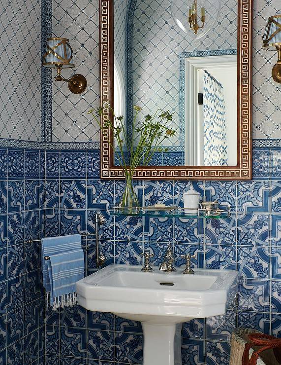 Mediterranean style bathroom features a gold Greek key mirror hung from a wall clad in mixed white and blue mosaic tiles between brass sconces and over a glass shelf. The shelf is fixed above a white porcelain pedestal sink vanity finished with a polished nickel faucet kit.