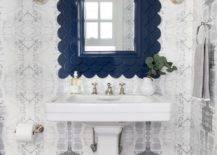 Chic white and navy blue bathroom showcases a navy blue scalloped mirror flanked by polished nickel sconces and hung from a wall clad in white and gray wallpaper over a white pedestal sink fixed to gray staggered floor tiles.