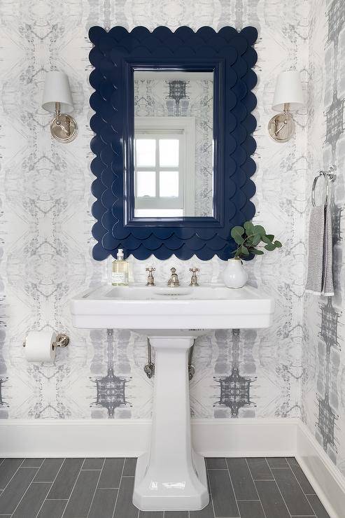 Chic white and navy blue bathroom showcases a navy blue scalloped mirror flanked by polished nickel sconces and hung from a wall clad in white and gray wallpaper over a white pedestal sink fixed to gray staggered floor tiles.