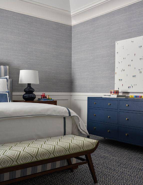 A canvas art piece hangs over a blue dresser accented with brass knobs and placed on blue and gray carpeting against a gray grasscloth clad wall finished with wainscoting. A bench with a green diamond print cushion is positioned at the foot of a gray striped bed accented with white and blue bedding lit by a blue double gourd lamp placed on a brown wood nightstand.