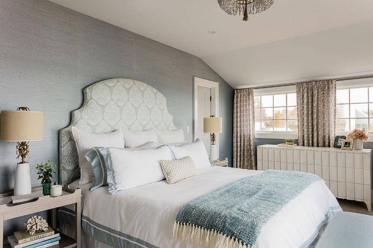 Gray tray nightstands topped with white and gold lamps flank a white and gray French headboard accenting a bed dressed in white and blue bedding draped in a blue herringbone throw blanket complementing blue grasscloth covered walls.