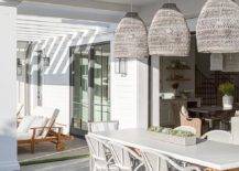 Placed on gray herringbone tiled pavers, white and gray bistro chairs and a gray wicker captain's chairs sit at a white and tan trestle table. The table is lit by three gray woven basket chandeliers hung from a white plank patio ceiling.