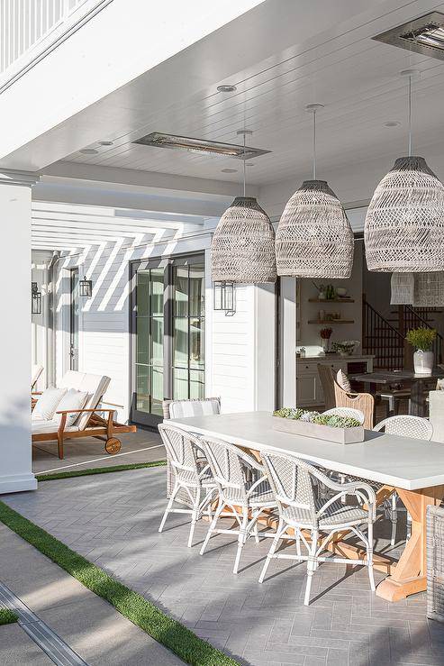 Placed on gray herringbone tiled pavers, white and gray bistro chairs and a gray wicker captain's chairs sit at a white and tan trestle table. The table is lit by three gray woven basket chandeliers hung from a white plank patio ceiling.