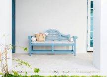 Outdoor cottage patio charmingly designed with a blue wooden bench with a light gray cushion. White exterior walls and lush greenery offer a streamlined template for added decor.