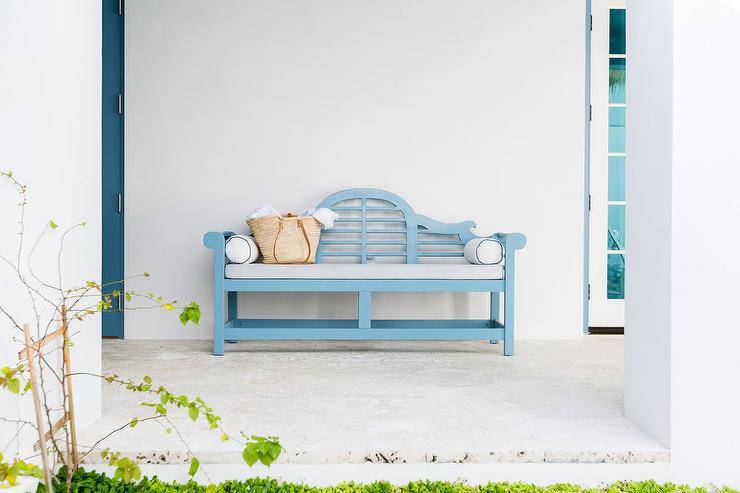 Outdoor cottage patio charmingly designed with a blue wooden bench with a light gray cushion. White exterior walls and lush greenery offer a streamlined template for added decor.