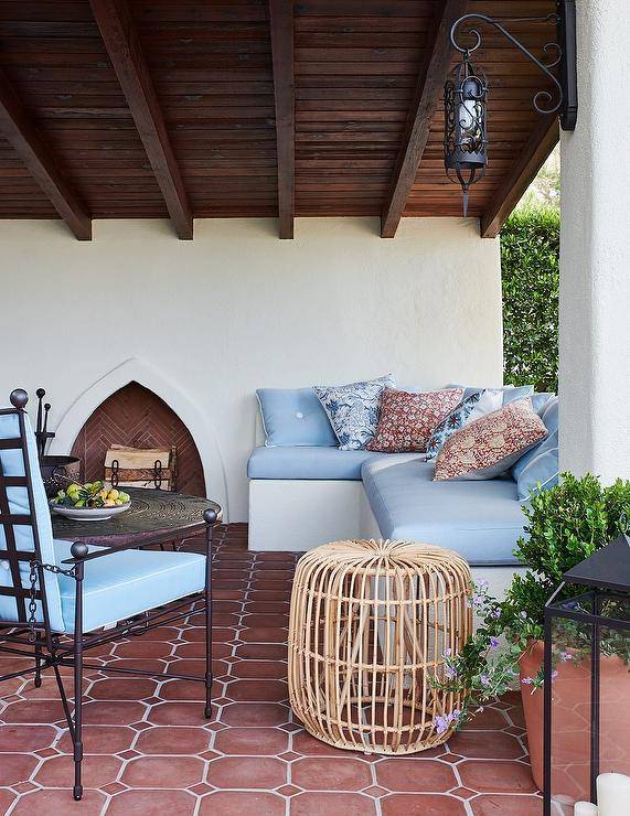 Stained wood plank ceiling is fitted over a patio boasting an l-shaped built-in sofa topped with a blue cushion and fixed against terracotta pavers beside a Moroccan silhouette fireplace.