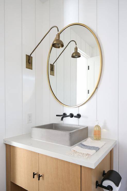 A gold vanity mirror is hung from a white vertical plank trim and lit by a brass swing arm sconce. Beneath the mirror, an oil rubbed bronze wall mount faucet is fitted above a concrete vessel sink mounted to a bronze sink vanity.