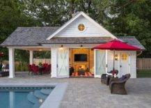 Cabin style pool house features double glass doors with rustic decor and a nautical interior design. Gray brick pavers finish the exterior patio floors surrounding a pool and a set of brown wicker chairs meeting red shade umbrella.