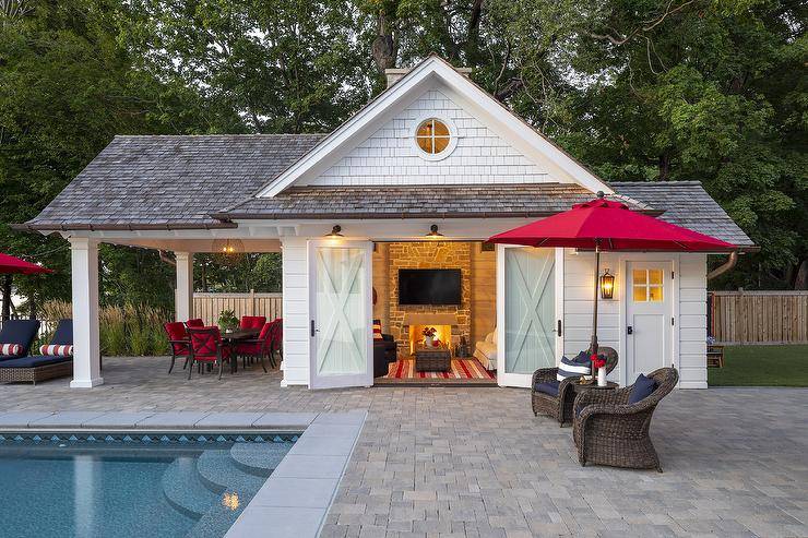 Cabin style pool house features double glass doors with rustic decor and a nautical interior design. Gray brick pavers finish the exterior patio floors surrounding a pool and a set of brown wicker chairs meeting red shade umbrella.