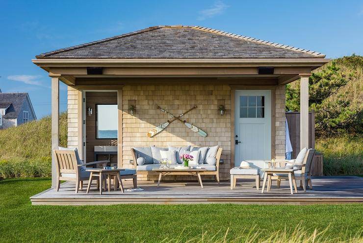 Relax on a cottage pool house deck with teak outdoor sofa, chairs and coffee table. Light brown shingles frame the deck displaying crossed canoe paddles flanked by a set of outdoor wall sconces. Light gray upholstery adds to a cozy patio decor accented with flower arrangements around a lush green outdoor setting.