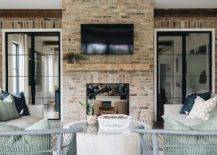 A flat panel TV is mounted over a rustic wooden mantel accenting a red brick fireplace fitted in a covered patio between bi-fold steel and glass doors. Gray rattan chairs sit on a beige striped rug facing the fireplace.