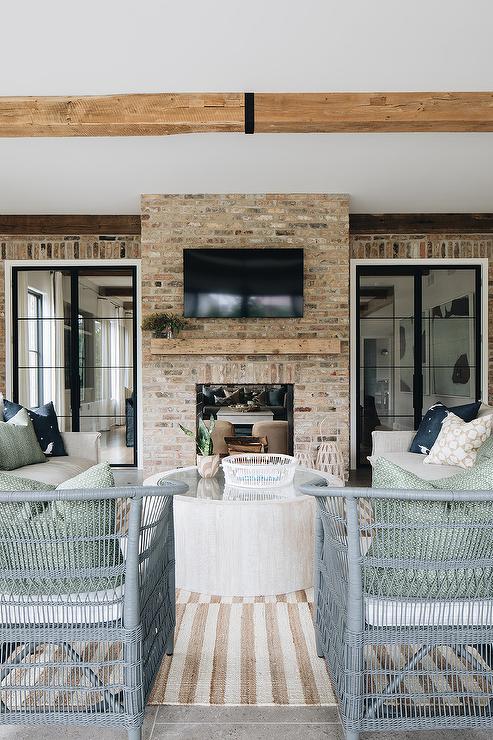 A flat panel TV is mounted over a rustic wooden mantel accenting a red brick fireplace fitted in a covered patio between bi-fold steel and glass doors. Gray rattan chairs sit on a beige striped rug facing the fireplace.
