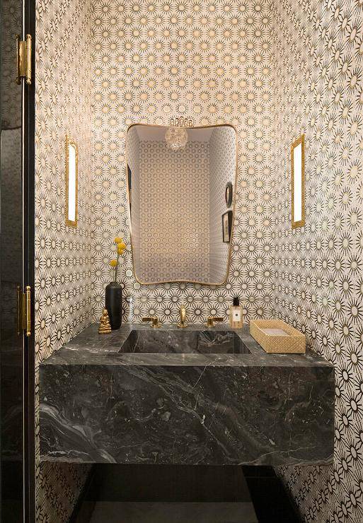 A brass mirror hangs from a wall clad in gold sunburst wallpaper over a black marble sink vanity with a brass faucet it.