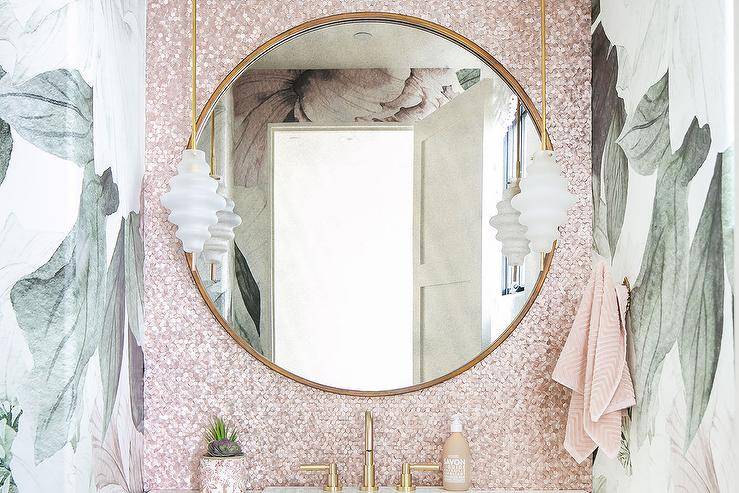 A round gold mirror hangs from pink iridescent backsplash tiles lit by brass and rippled glass mini pendants fixed over a brass faucet kit.