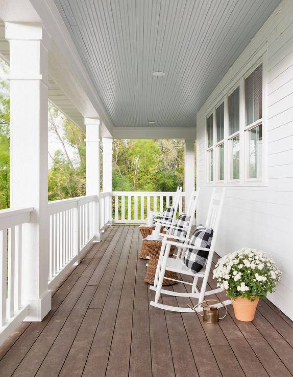 Wraparound porch furnished with white rocking chairs and black buffalo check pillows. A blue porch ceiling, woven accent tables, and potted plants deliver a charming design adding to the homes curb appeal.
