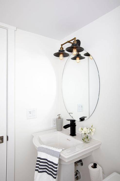 A frameless oval family mirror is lit by an oil rubbed bronze 2-light vintage barn sconce and fixed over a curved pedestal sink with a matte black faucet kit.