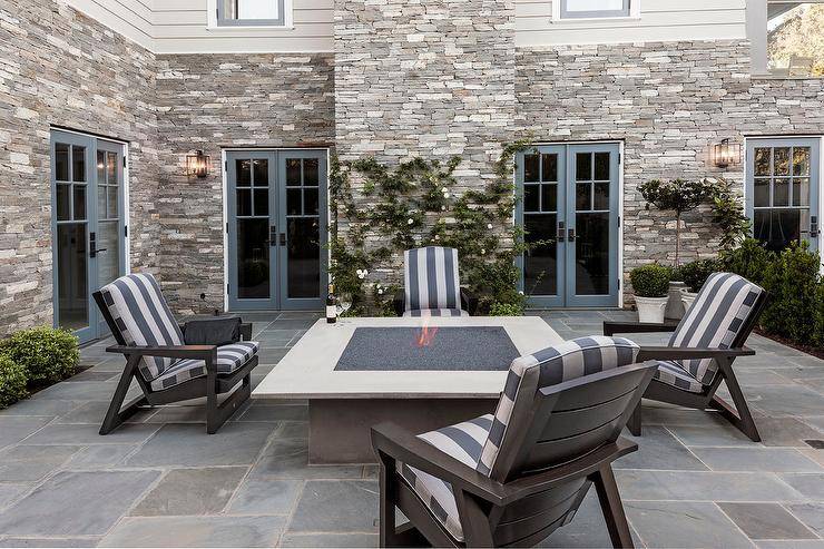 Blue framed french doors lead to a gorgeous patio boasting an oversized square concrete gas fire pit surrounded by dark gray Adirondack chairs topped with white and blue striped seat cushions and sat upon shrub lined stunning slate pavers.