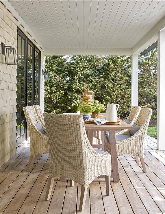 Beneath a covered porch, a gray wash dining table is surrounded by wicker dining chairs.