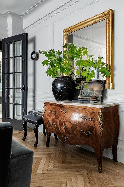 Wood herringbone pattern flooring leads to a marble top bombay chest with gold baroque mirror, and light gray walls with light gray wainscoting in this transitional entryway.